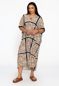 Robe longue et ample en marron et blanc avec des motifs géométriques. Présente un décolleté en V et une boucle décorative. Assortie à des sandales marron.