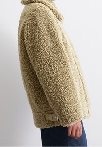 Tann, fluffig fårskinnsjacka med texturerad yta, bred krage och sidodetalj i metall. Designen har en avslappnad passform och långa ärmar.