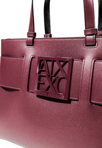Burgundowa syntetyczna torebka z teksturowanym wykończeniem, z czarną skórzaną rączką i wyraźnym wytłoczonym logo na przodzie.