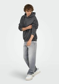 Joven en un hoodie gris oscuro arremangándose las mangas, vistiendo jeans gris claro y zapatillas blancas, de pie contra un fondo claro y sencillo.