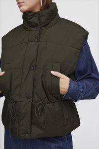 Pehmea oliivroheline vest kõrge krae, nööbivaba sulgemise, küljetaskute ja reguleeritava tõmbluku seelikuosa. All on teksajakk.