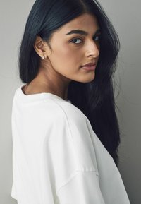 Hvid oversized sweatshirt med rund halsudskæring, sat mod en neutral baggrund. Langt, glat sort hår og en simpel sølvørering er synlig.