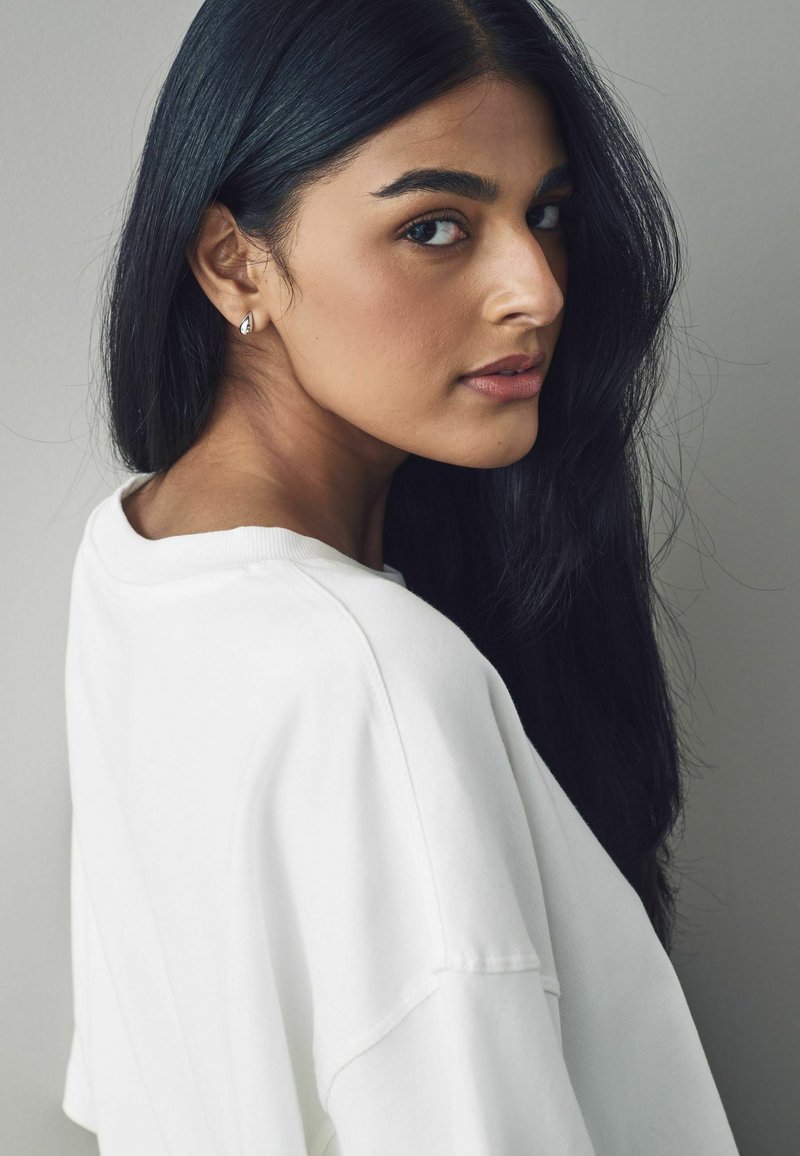 Hvid oversized sweatshirt med rund halsudskæring, sat mod en neutral baggrund. Langt, glat sort hår og en simpel sølvørering er synlig.