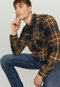 Camisa de franela a cuadros marrón y azul marino con cuello abotonado, dos bolsillos en el pecho y mangas largas, combinada con jeans de mezclilla azules.