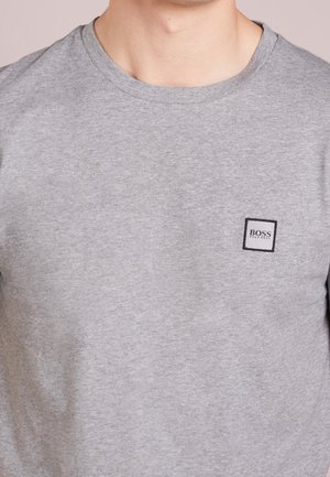 Męski tors w jasnoszarej koszulce z okrągłym dekoltem, z małą prostokątną naszywką z logo "BOSS HUGO BOSS" na lewej piersi.