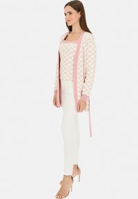 Jasnoróżowy kardigan z geometrycznym wzorem na dopasowanej bluzce, zestawiony z białymi obcisłymi spodniami i beżowymi szpilkami; gładka tekstura tkaniny.