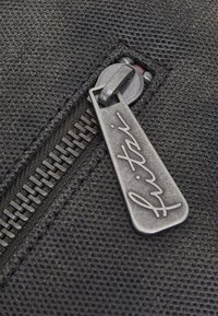 Cerniera in tessuto nero testurizzato con cursore metallico dotato di targhetta incisa con firma. I denti della cerniera sono visibili accanto al cursore.