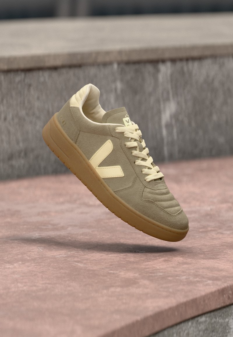 Μπεζ σουέτ αθλητικό παπούτσι με λαστιχένια σόλα και εκρού κορδόνια, με το λογότυπο "V" στην πλευρά, αιωρούμενο πάνω από ροζ λιθόστρωτο με γκρι πέτρινο υπόβαθρο.