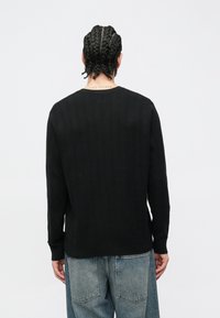 Μαύρο ριμπ πλεκτό πουλόβερ με στρογγυλή λαιμόκοψη, μακριά μανίκια και ελαφρώς εφαρμοστή μέση. Φορεμένο με ανοιχτόχρωμα τζιν παντελόνια.