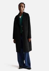 Cappotto di lana nero con rever a V, due tasche anteriori e chiusura frontale con bottoni, indossato sopra una maglia verde e pantaloni in denim a gamba larga.