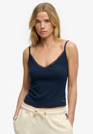 Femme blonde portant un haut sans manches bleu marine avec bordure en dentelle et un pantalon crème à cordon, debout les mains dans les poches sur fond blanc.