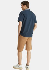 Polo shirt blu navy con bordo bianco, pantaloncini khaki e sneakers alte bianche. Il modello è in piedi con le mani in tasca, visto da dietro.