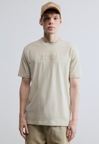 Bézs pamut póló dombornyomott "BOSS" logóval a mellkason, kerek nyakkivágással és rövid ujjú. Egyenlő színű bézs sapkával párosítva.