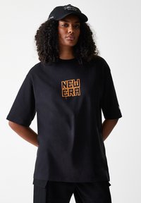 T-shirt nera oversize con design arancione "NEW ERA" sulla parte anteriore, texture morbida e maniche corte. Abbinata a un cappellino nero.