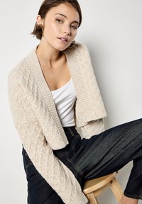 Cárdigan beige de punto grueso con cuello acanalado, abertura frontal y mangas largas, combinado con una camiseta blanca y pantalones vaqueros oscuros.