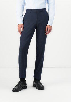 Homem vestindo calças de fato azul marinho sob medida, sapatos de dress pretos e uma camisa de dress azul clara, de pé contra um fundo branco.