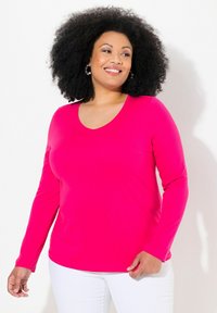 Růžové tričko s dlouhým rukávem a výstřihem do V vyrobené z hladkého materiálu, s přiléhavým střihem, v kombinaci s bílými kalhotami.