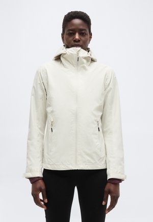 Persoană cu păr scurt purtând o jachetă albă cu glugă și fermoar și pantaloni negri, pe un fundal simplu, deschis la culoare.