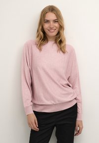 Rózsaszín, laza kötésű pulóver hosszú ujjal, kerek nyakkivágással, bordázott mandzsettával és aljjal, fekete nadrággal párosítva.