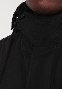 Nahaufnahme einer Person, die eine schwarze Jacke mit hohem Kragen, Knopf und Zugband trägt, vor einem schlichten Hintergrund.