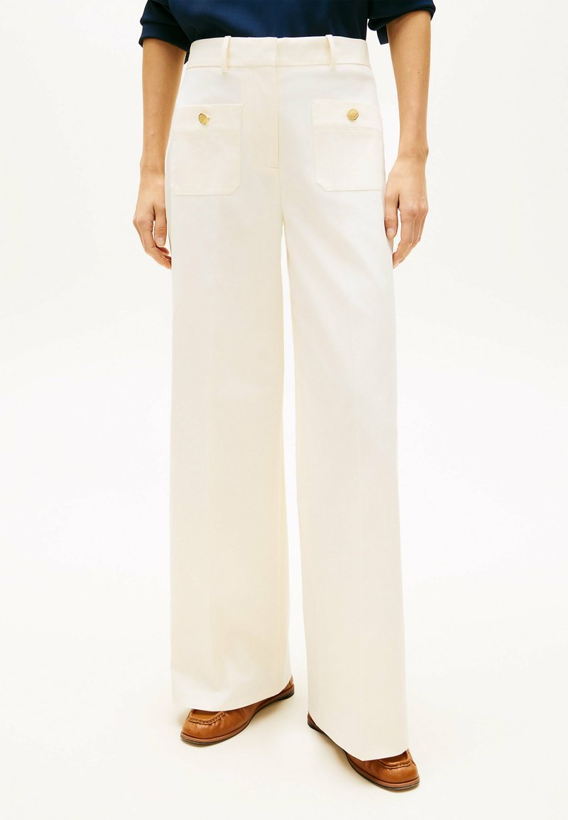 Vrouw die crèmekleurige, hoog getailleerde, wijde broek draagt met voorzakken en bruine loafers, op een witte achtergrond.