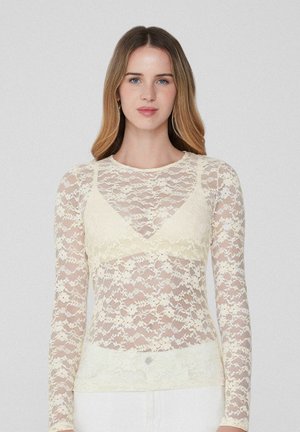 Femme aux longs cheveux châtain clair portant un haut à manches longues en dentelle florale crème sur un bralette assorti, se tenant devant un fond uni.