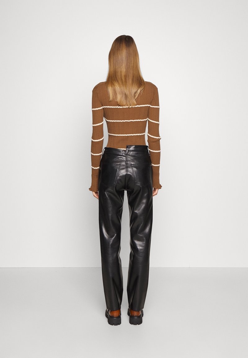 Femme debout de dos portant un haut à manches longues marron rayé et un pantalon en cuir noir avec des chaussures noires épaisses.