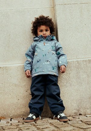 Enfant aux cheveux bouclés portant une veste bleue avec des dessins de véhicules, un pantalon marine et des baskets, debout contre un mur de pierre sur des pavés.