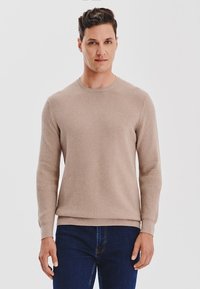 Beżowy sweter dzianinowy o teksturowanym wzorze, z okrągłym dekoltem i długimi rękawami. Noszony z ciemnoniebieskimi dżinsami. Gładki materiał przypominający wełnę.