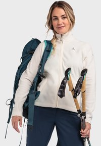 Leichte weiße Fleecejacke mit hohem Kragen, blauen Rucksack und Trekkingstöcke mit Korkgriffen und schwarzen Schlaufen.