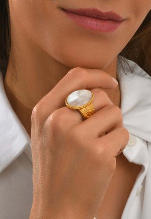 Gros plan d'une main près du visage portant une bague en or avec une grande pierre blanche ovale, sur fond de chemise blanche à col.