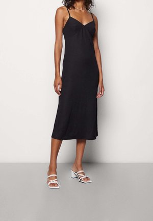 Kobieta w czarnej, midi sukience bez rękawów z cienkimi ramiączkami oraz białymi sandałami na blokowym obcasie, stojąca na tle gładkiej ściany.