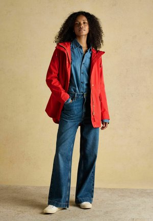 Kobieta o kręconych włosach, ubrana w czerwoną kurtkę na dżinsowej koszuli i szerokich spodniach dżinsowych, stojąca z jedną ręką w kieszeni.
