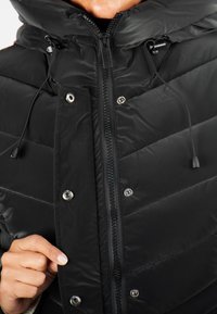 Veste matelassée noire avec un col haut, un design matelassé diagonal, des boutons-pression argentés et un cordon de serrage pour un ajustement réglable. Fermeture éclair.