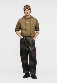 Jeune homme portant un bonnet brun rayé, un sweat à capuche vert olive avec logo, des manches longues rayées, un pantalon cargo noir et brun, et des chaussures noires.