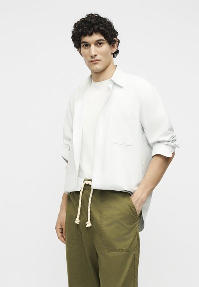 Νέος άνδρας με σγουρά σκούρα μαλλιά, φοράει λευκό πουκάμισο πάνω από λευκό μπλουζάκι, ελιτί πράσινο παντελόνι με λευκή ζώνη σχοινί, στέκεται με το ένα χέρι στην τσέπη.