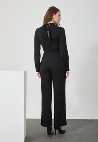 Combinaison noire avec des manches longues, un corsage ajusté et des jambes larges. Présente un dos en forme de clef et des liens au cou. Texture lisse et design ajusté.