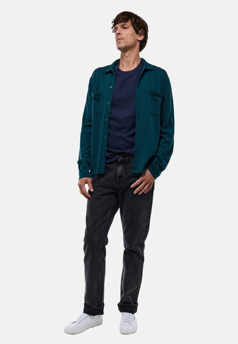 Chemise bleu canard à col boutonné avec deux poches à l'avant, portée sur un t-shirt bleu marine, associée à un jean foncé et des baskets blanches, offrant une coupe décontractée.