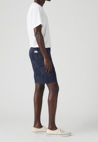 Hombre con camiseta blanca, pantalones cortos con estampado en azul marino y zapatillas de lona color crema de pie de lado sobre un fondo liso.