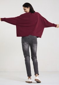 Burgunderfarbener Over-size-Pullover mit weiten Ärmeln, geripptem Saum, kombiniert mit enganliegenden grauen Jeans und weißen Sneakern mit braunen Akzenten.