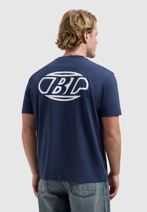 Man met blond haar die een marineblauw T-shirt draagt met een groot wit BP-logo op de achterkant, gecombineerd met lichtblauwe spijkerbroek.