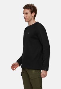 Man wearing black long-sleeve textured sweater and olive green cargo pants standing against a white background, facing left.
