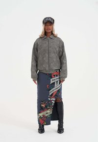 Giacca in finta pelle grigia con colletto, abbinata a una gonna blu con una grafica ispirata ai tatuaggi e uno spacco laterale. Stivali neri fino al ginocchio.