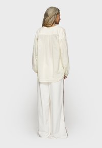 Maglietta a maniche lunghe color crema con vestibilità ampia, caratterizzata da dettagli arricciati sul retro, abbinata a pantaloni bianchi a gamba larga con strisce laterali marroni.