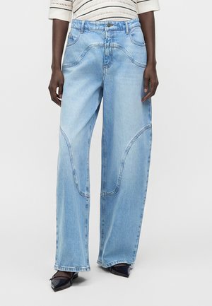 Vaqueros de pierna ancha azul claro con detalles de costuras curvas, combinados con zapatos negros puntiagudos y una camiseta de rayas de manga larga.