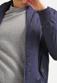 Námořnická modrá lehká bunda s hladkým povrchem, s předním zapínáním na knoflíky a elastickými manžetami, nošená přes šedou mikinu s kulatým výstřihem.