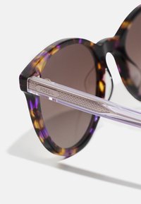 Runde Sonnenbrillen mit einem lebhaften Schildpatt-Herz in lila und amberfarbenen Tönen. Mit transparenten Akzenten und braun getönten Gläsern.