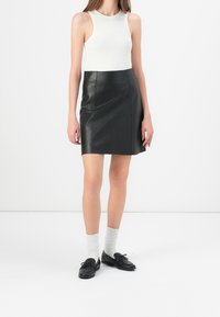 Mini gonna nera in pelle a campana, top bianco a coste senza maniche, scarpe nere con fiocchi e calze bianche. Sfondo neutro.