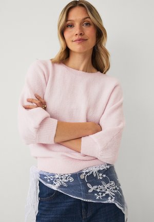 Blondynka z założonymi ramionami ma na sobie jasnoróżowy sweter i niebieskie dżinsy, a wokół talii związany jest biały koronkowy szal w kwiaty.