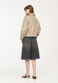 Giacca in suede beige con colletto alto, abbinata a pantaloni shorts in denim scuro lunghezza ginocchio con bordi sfrangiati, e sandali marroni con laccetto.